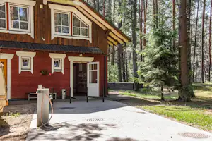 Holz-Ferienhaus mit Terrasse im Wald, Eingangstür, Markise, Tisch und Gasgrill auf Betonfläche.