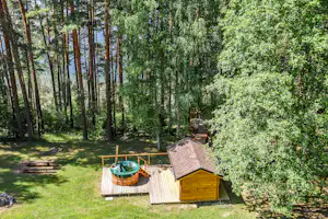 Holzterrasse mit Gartenhütte und Schubkarre, dahinter ein Weg durch den Wald mit Blick auf den See.