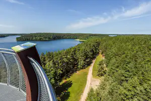 Blick vom Aussichtsturm auf einen See, Wald und einen Weg im Campinggelände, im Vordergrund Geländer und Infotafel.