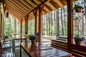 Holzveranda mit gedecktem Dach, Tischen und Bänken, Blick auf den Wald mit hohen Bäumen und Kiesweg.