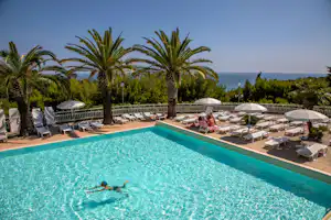 Schwimmbecken mit Liegestühlen, Sonnenschirmen und Palmen am Campingplatz, im Hintergrund das Meer.