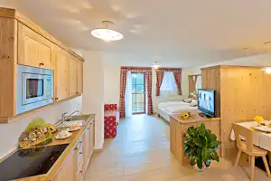 Wohnbereich mit Küchenzeile aus Holz, Esstisch und Fernseher, daneben ein Bett und Fenster mit roten Vorhängen.