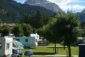Campingplatz mit Wohnmobilen und Zelten, Rasenfläche und Bäumen vor hohen Bergen im Hintergrund.
