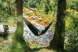 Grüne Hängematte zwischen Bäumen am Flussufer, im Hintergrund Wiese und Wasserlauf im Wald.