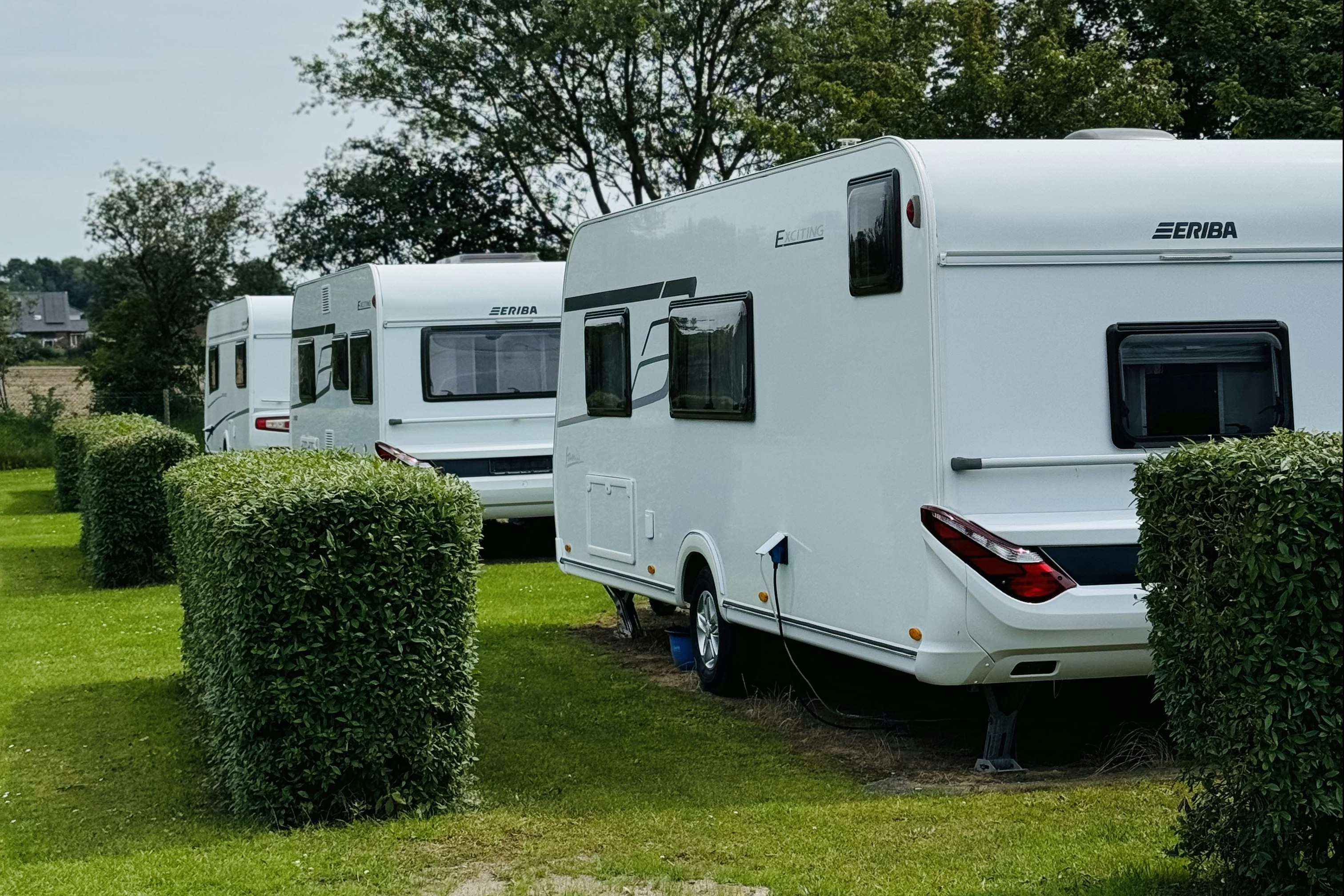 Wohnwagen auf einem Campingplatz, mit Hecken und Rasenfläche, im Hintergrund weitere Wohnwagen und Bäume.