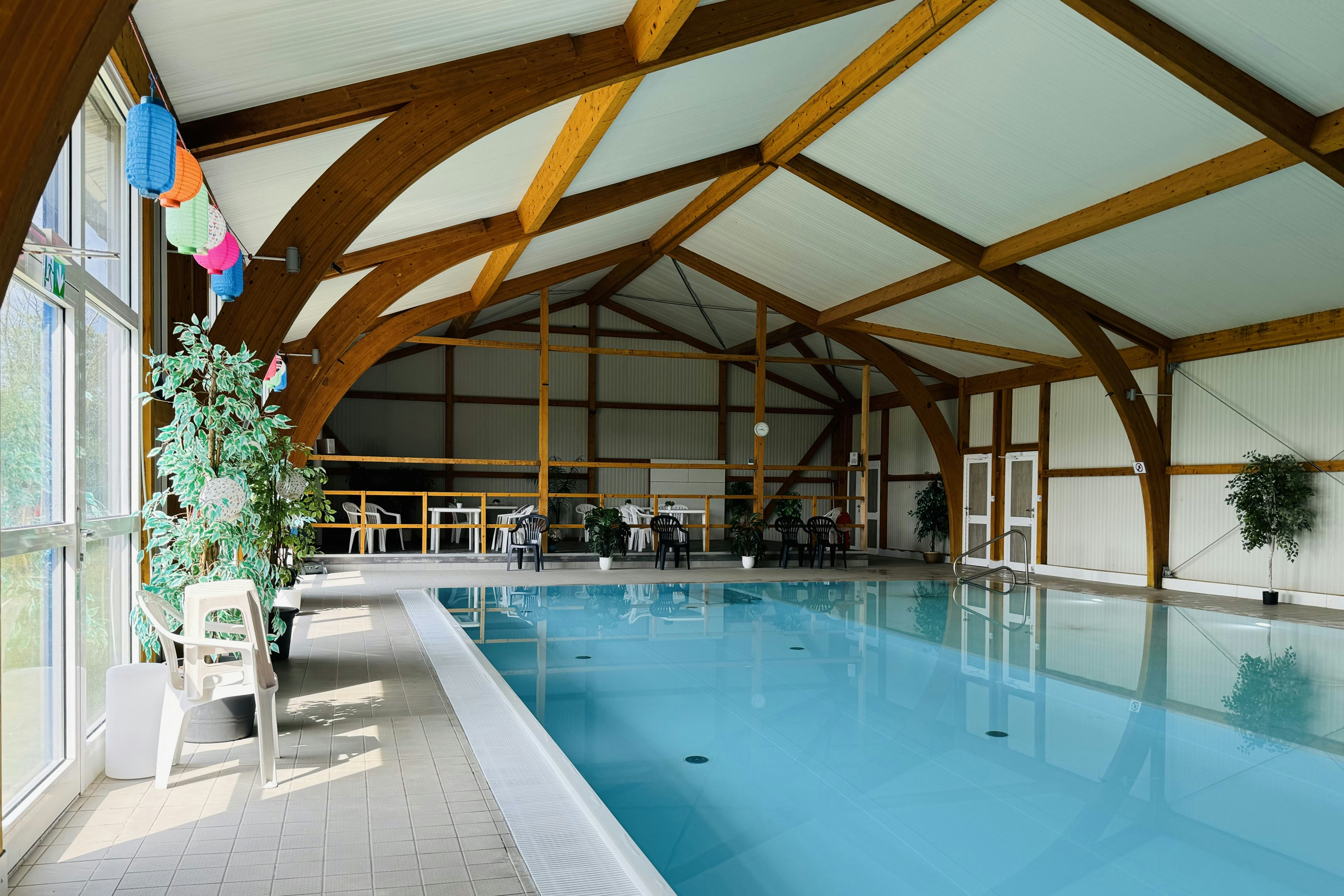 Innenbereich mit beheiztem Schwimmbecken unter Holzgewölbe, Liegen und Sitzgelegenheiten, große Fenster und Pflanzen im Hintergrund.