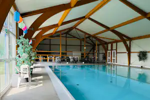 Innenbereich mit beheiztem Schwimmbecken unter Holzgewölbe, Liegen und Sitzgelegenheiten, große Fenster und Pflanzen im Hintergrund.