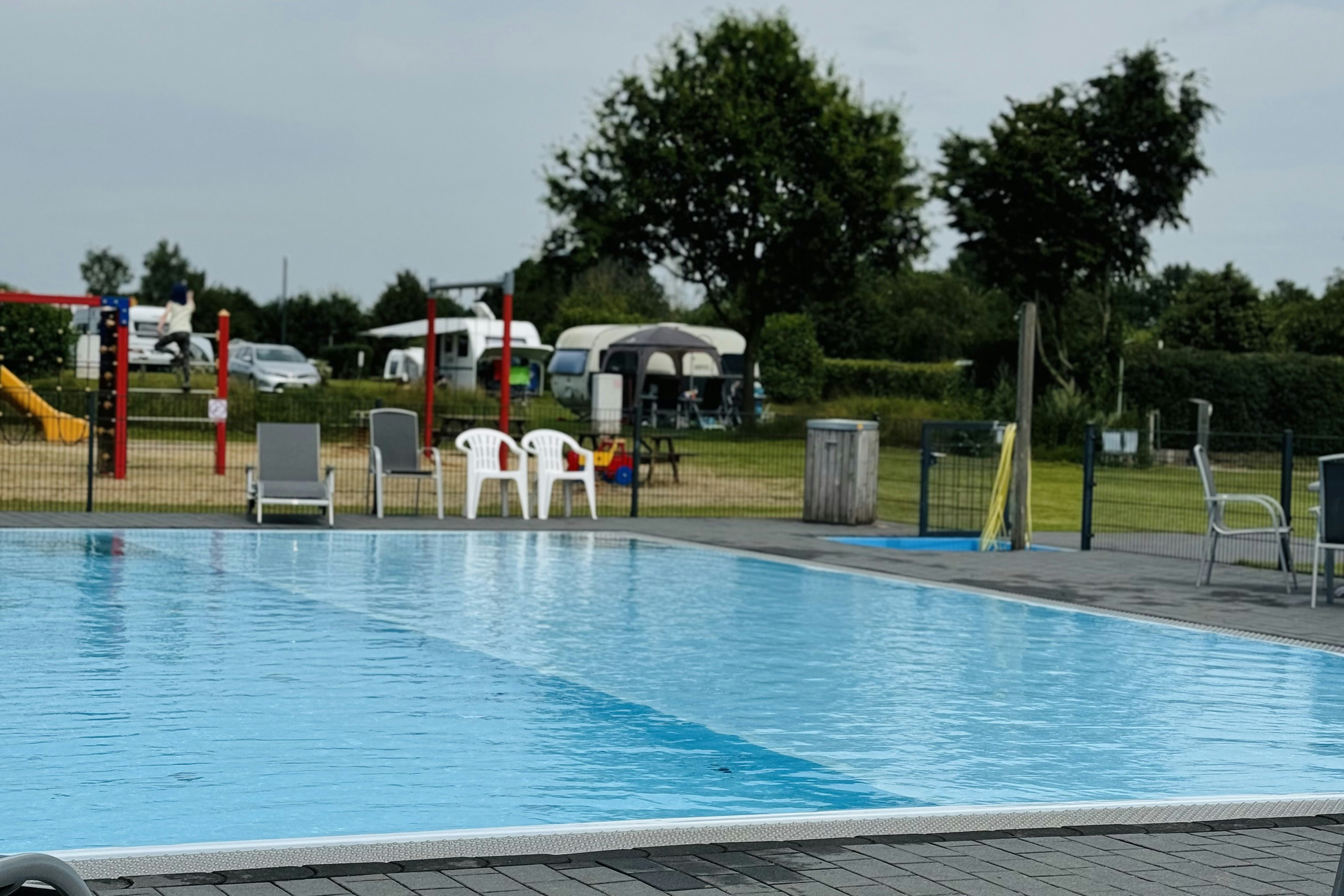 Schwimmbecken mit blauem Wasser, daneben Liegen und Absperrpfosten auf dem Campingplatz, im Hintergrund Wohnwagen und Bäume.