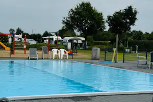 Schwimmbecken mit blauem Wasser, daneben Liegen und Absperrpfosten auf dem Campingplatz, im Hintergrund Wohnwagen und Bäume.