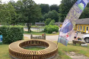 Campingplatz mit gelben Ferienhäusern, Gartenweg und großer Werbefahne vor einem Holzfeuerkreis, umgeben von Bäumen.