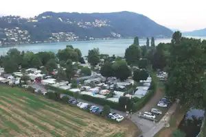 Campingplatz am See mit vielen Stellplätzen, Wohnwagen und Zelten, umgeben von Bäumen und Blick auf die Berge.
