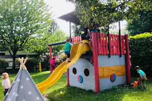 Spielgerät mit gelber Rutsche und rotem Holzrahmen im grünen Campinggarten, Kinder klettern und spielen.