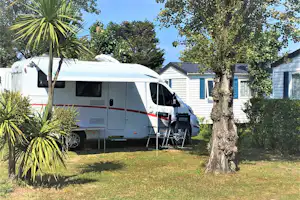 Wohnmobil mit Markise und Sitzgruppe vor weißen Mobilheimen, umgeben von Bäumen und Palmen im Campinggarten.