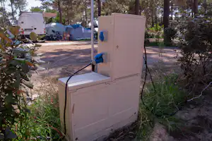 Stromanschluss-Box auf einem Campingplatz-Stellplatz, im Hintergrund Wohnwagen und Zelte zwischen Bäumen.