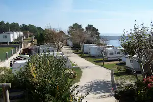 Campingplatz mit Wohnwagen entlang eines Weges, Hecken und Bäumen, im Hintergrund das Meer.