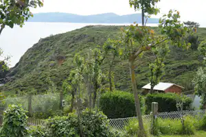 Campingzaun und Hecken vor einem kleinen Holzhaus, dahinter grüner Hügel und See mit Bergen in der Ferne.