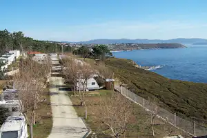 Campingplatz am Meer: Wohnwagen und Stellplätze auf einem Hangweg, Bäume und Hecken entlang der Küste, Blick aufs Wasser.