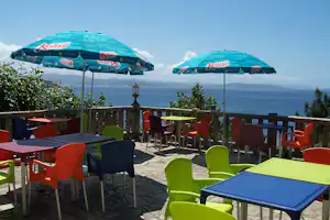 Terrasse mit bunten Stühlen und Tischen, zwei Sonnenschirmen und Blick auf das Meer und die Berge in der Ferne.