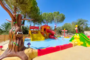 Spielbereich mit Wasserrutsche und Hüpfburg, im Vordergrund eine große Holzfigur, daneben Bäume und ein roter Zaun.