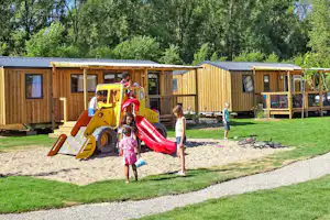 Spielplatz am Campingplatz mit gelber Rutsche, Kindern und buntem Spielgerät vor Holzunterkünften und Rasen.