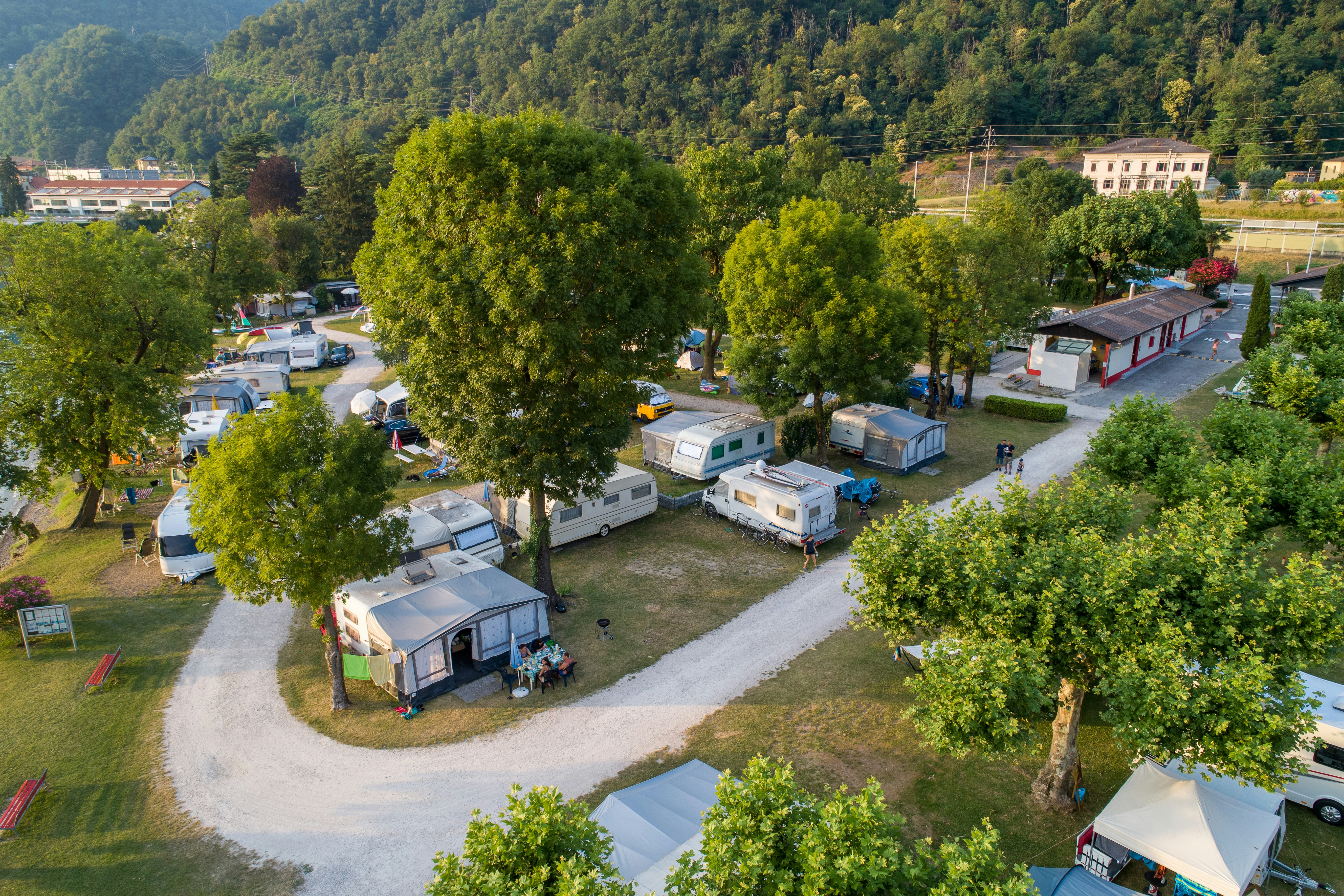 Luftaufnahme eines Campingplatzes mit Wohnwagen und Zelten, gepflasterten Wegen, Bäumen und einem Gebäude am Rand.