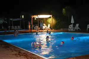 Beleuchteter Camping-Pool bei Nacht mit Kindern im Wasser und Liegestühlen auf der Terrasse unter einem Sonnensegel.