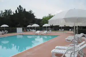 Poolbereich mit Sonnenschirmen, Liegestühlen und gepflasterter Terrasse am Campingplatz, im Hintergrund Hecken und Bäume.