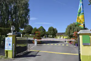 Einfahrt zum Campingplatz mit Schranke, Flagge und Blumenbeeten, dahinter Bungalows und Bäume unter blauem Himmel.