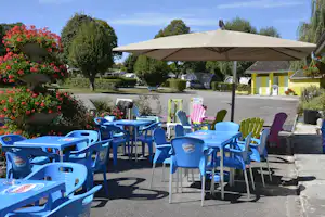 Camping-Terrasse mit Sonnenschirm, bunten Plastikstühlen und -tischen, Hecken, Blumenbeeten und Stellplätzen mit Autos im Hintergrund.