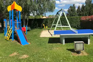 Spielplatz mit blauer Rutsche, Schaukeln und Sandkasten auf einer Wiese zwischen Bäumen am Campingplatz.