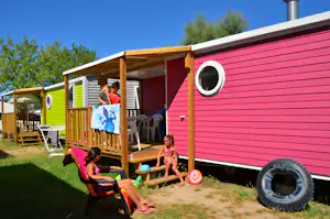Buntes Mobilheim mit Holzveranda, Sitzgelegenheiten und Wäscheleine; Kinder spielen im Vordergrund, daneben ein Reifen.
