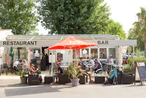 Restaurant und Bar auf der Camping-Terrasse mit Sonnenschirmen, Tischen und Stühlen vor großen Fenstern.
