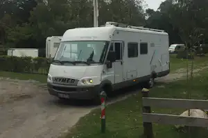 Weißes Wohnmobil auf einem Campingplatz-Parkplatz, mit Schranke und Weg, umgeben von Bäumen und Rasen.