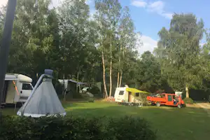 Zelt auf einem Campingplatz-Rasen, daneben Wohnwagen und Wohnmobil, umgeben von hohen Bäumen und einem gepflegten Weg.
