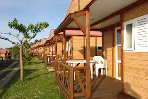 Holzterrasse am Bungalow mit weißen Stühlen, Geländer und Blick in den Garten mit Bäumen und Rasen.