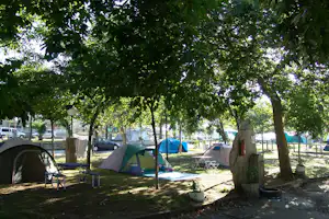 Zelte auf einem Campingplatz unter Bäumen, mit Campingstühlen und Stellplätzen, im Hintergrund Fahrzeuge und weiße Zeltplanen.