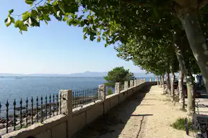 Uferpromenade am See mit Steinmauer, Bäumen und Schatten, im Hintergrund Wasser und entfernte Hügel.