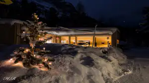 Beleuchtete Campinghütte im Schnee mit Weihnachtslichtern, Holzfassade und großen Fenstern, im Hintergrund dunkle Berge und Bäume.