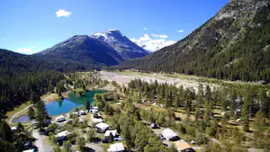 Campingplatz im Alpen-Tal mit Wohnmobilen und Zelten am türkisfarbenen See, umgeben von Wäldern und Bergen.