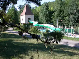 Weg im Campinggelände mit Olivenbaum, kleiner Kapelle und Pool hinter einem grünen Zaun, umgeben von Bäumen und Sträuchern.