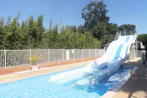 Camping-Pool mit blauer Rutsche, Sonnenterrasse und Zaun, umgeben von Bäumen und Sträuchern.