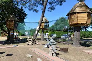 Holz-Spielhütten auf dem Campingplatz, mit Zaunnetz, Sitzbank und Spielbereich am Boden, im Schatten der Bäume.
