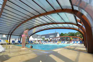 Innenpool mit Holzbogen, Becken und Liegestühlen unter Glasdach, im Hintergrund Gebäude und Bäume.