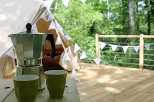 Camping-Tisch mit Kaffeekanne und Bechern vor Zelten, Holzterrasse und Bäumen im Hintergrund.
