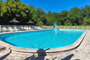 Campingpool mit blauem Wasser, Rutsche und weißen Liegen am Rand, umgeben von Bäumen und Zaun.