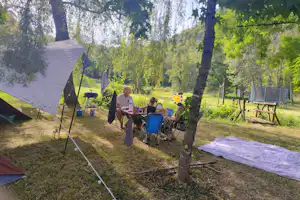 Campingplatz mit Zelt, Tisch und Stühlen im Grünen, daneben weitere Campingausrüstung zwischen Bäumen.