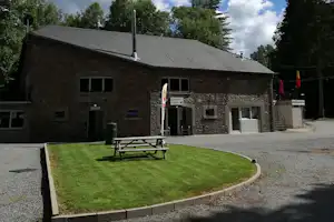 Campinggebäude aus Stein mit Eingang, davor ein gepflegter Rasen und eine Bank auf dem Kiesweg.