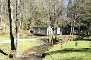 Campingplatz mit kleinen Gebäuden und Wohnwagen, Bachlauf durch die Wiese, umgeben von Bäumen im Frühling.
