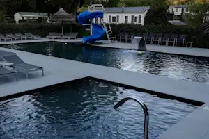 Schwimmbecken mit Wasserrutsche, Liegen am Beckenrand und weißen Gebäuden im Hintergrund auf dem Campingplatz.
