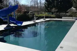 Blauer Pool mit Rutsche und Liegen auf einer hellen Terrasse, umgeben von Bäumen und Hecken.